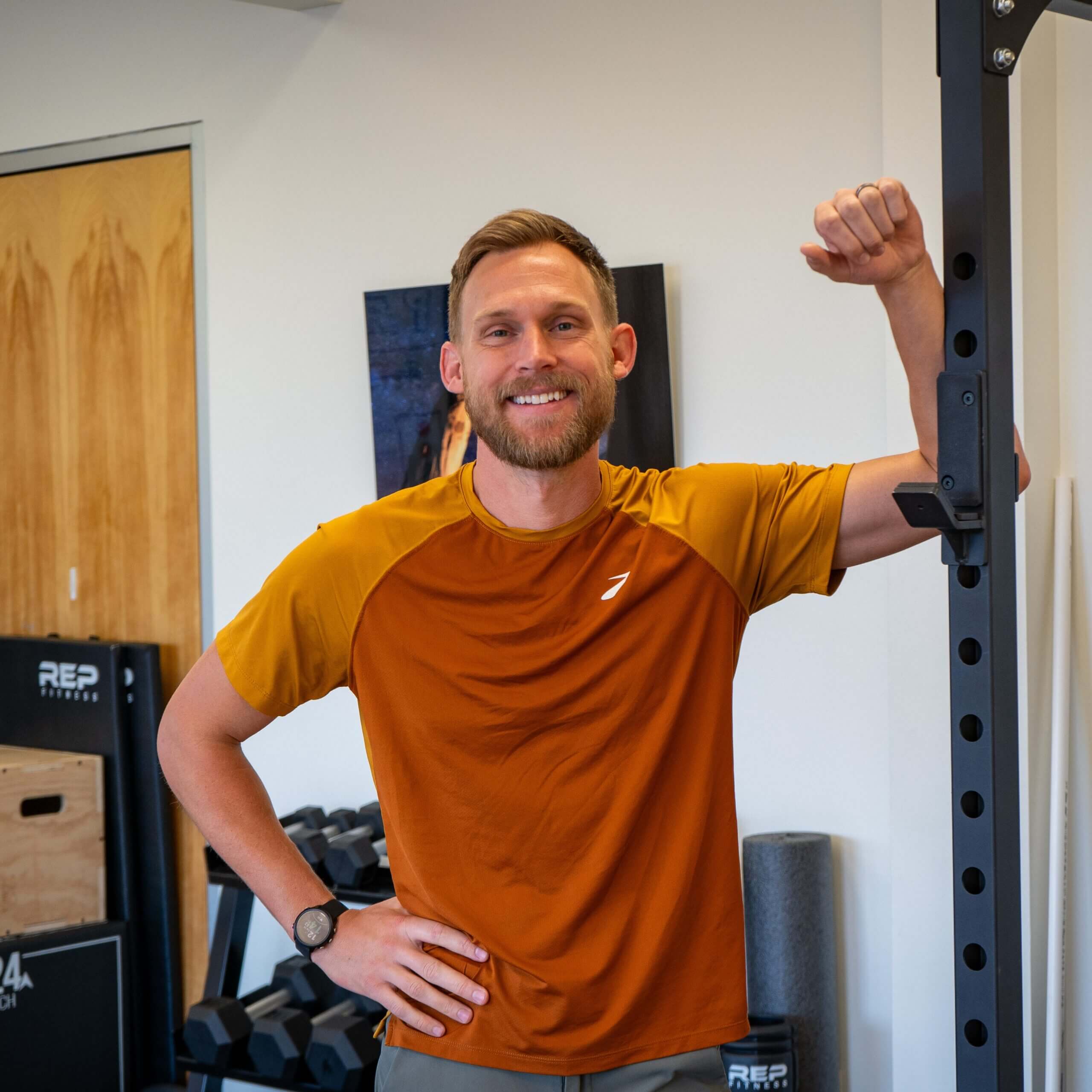 A smiling man in a mustard-yellow t-shirt stands beside physical therapy equipment, raising one arm in a flexed position, showing a casual and friendly demeanor in a fitness environment.