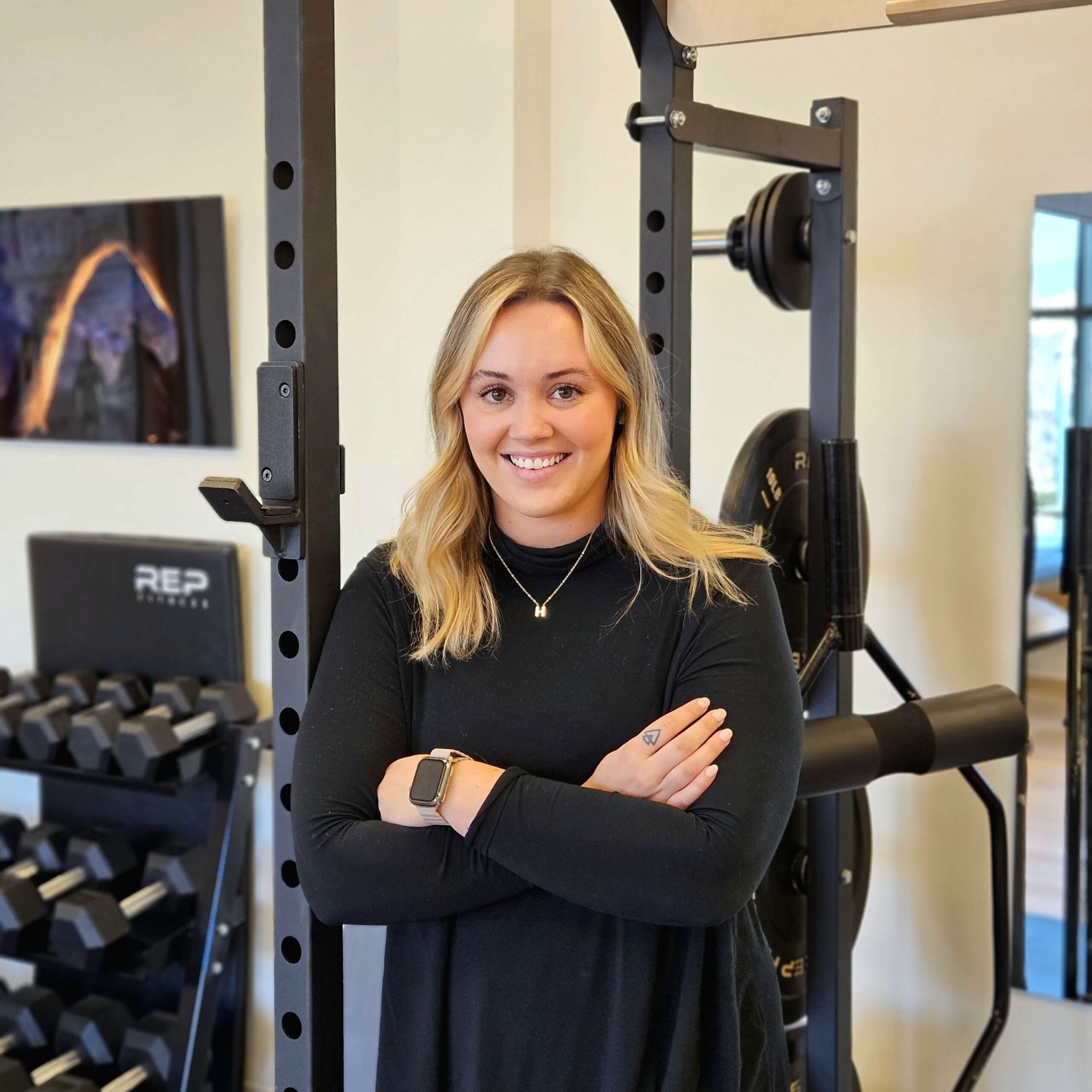 A woman with blonde hair smiling in a physical therapy gym, standing with her arms crossed in front of a weight rack. She is wearing a black long-sleeve shirt and a watch.