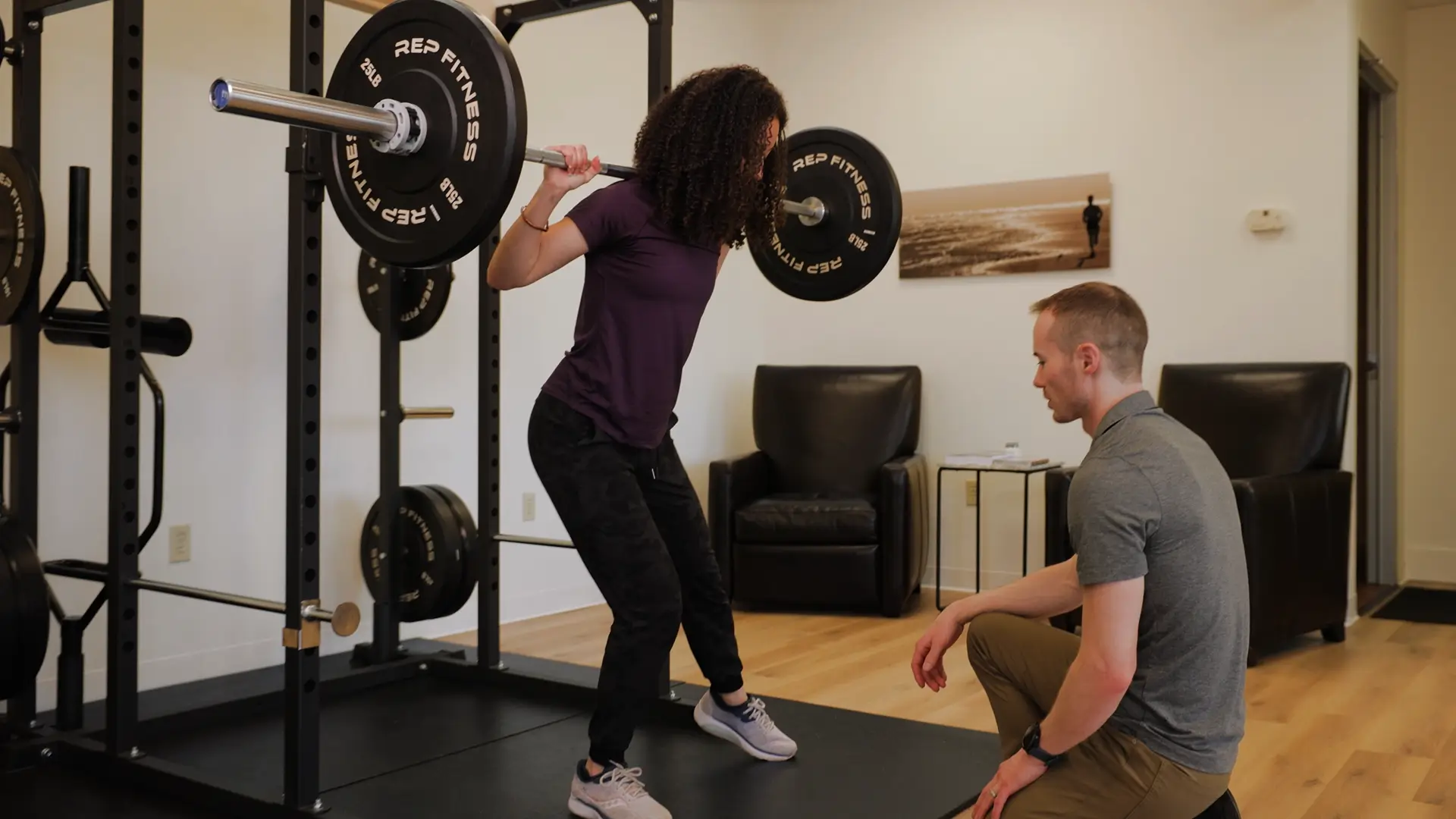 A person with curly hair performs a squat with a barbell on their shoulders in a gym, under the watchful eye of a seated physical therapy trainer nearby.