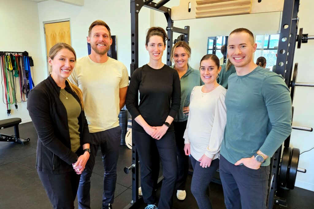 Group of six adults, smiling and posing together in a physical therapy gym with exercise equipment in the background. They are dressed in casual workout attire.
