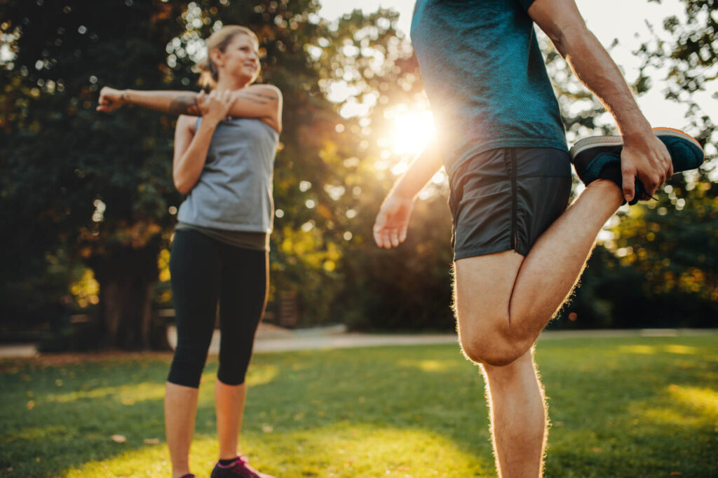Two people engaging in physical therapy in a sunny park, with a focus on a man stretching his leg and a woman in the background performing a torso twist. Sunlight adds a warm glow to the serene