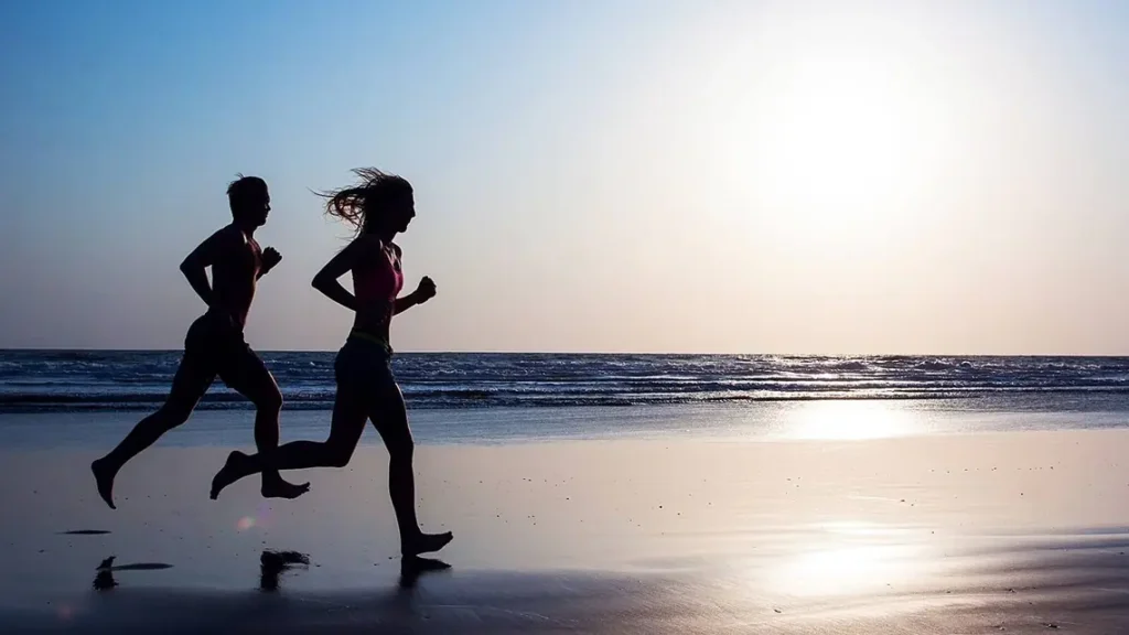 Silhouettes of a man and a woman running on a beach at sunset, with a bright sky and calm sea in the background.