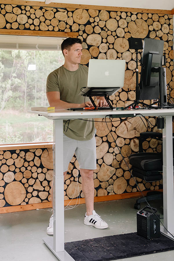 A man stands at an adjustable desk in a room with walls lined with logs, working on a laptop. He is dressed casually and appears focused on his screen. Nearby, physical therapy equipment suggests a multif
